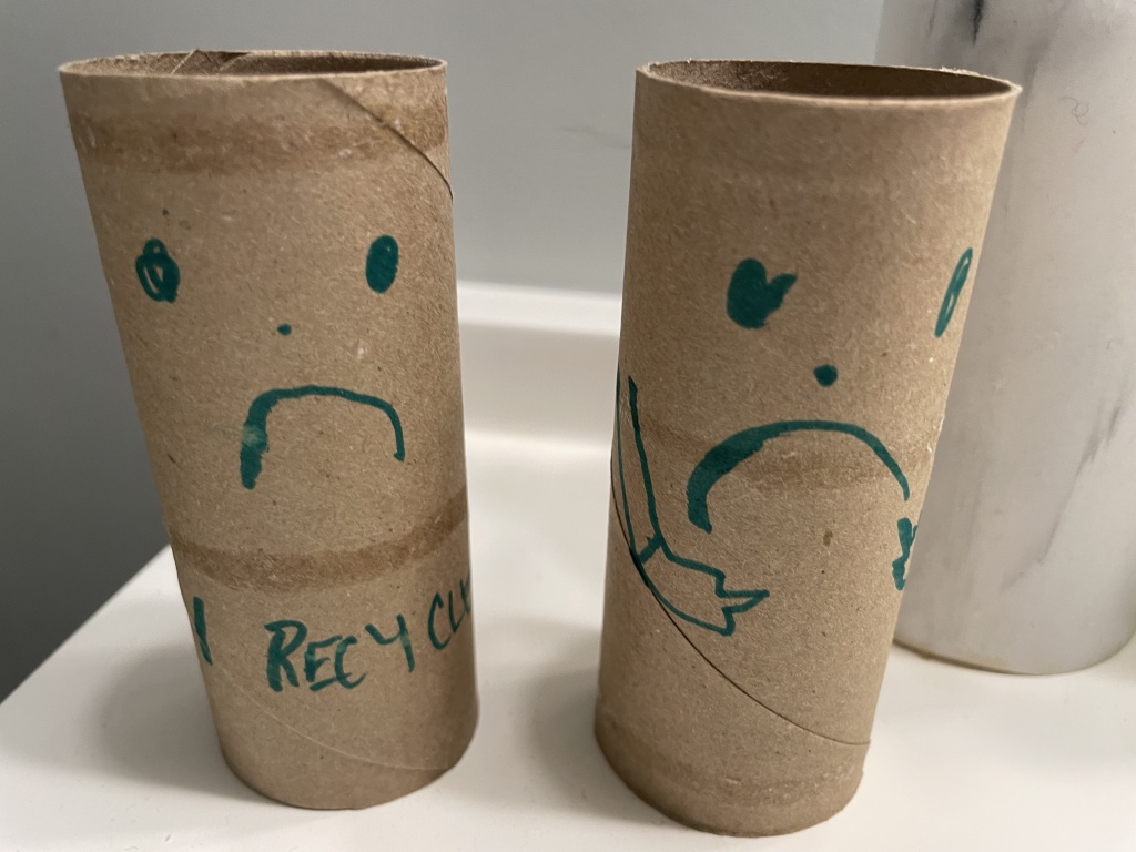 Two toilet paper rolls with faces and the words recycle me on them