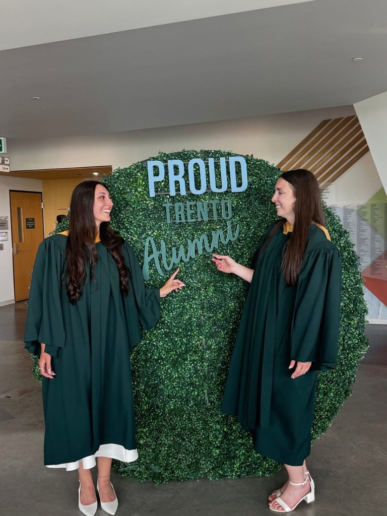 Two new Trent grads standing in front of an alumni sign in their gowns